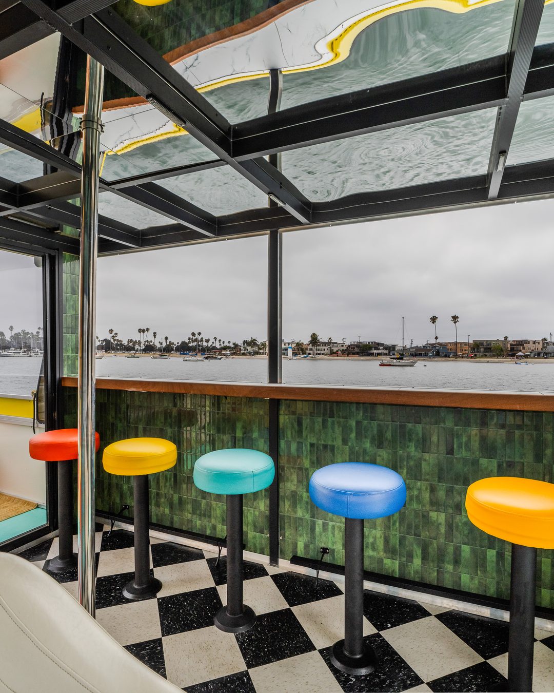 Disco Boat 5-seat rainbow bar — jewel-tone stools at the green-tile bartop, bay views all around, checkerboard floor