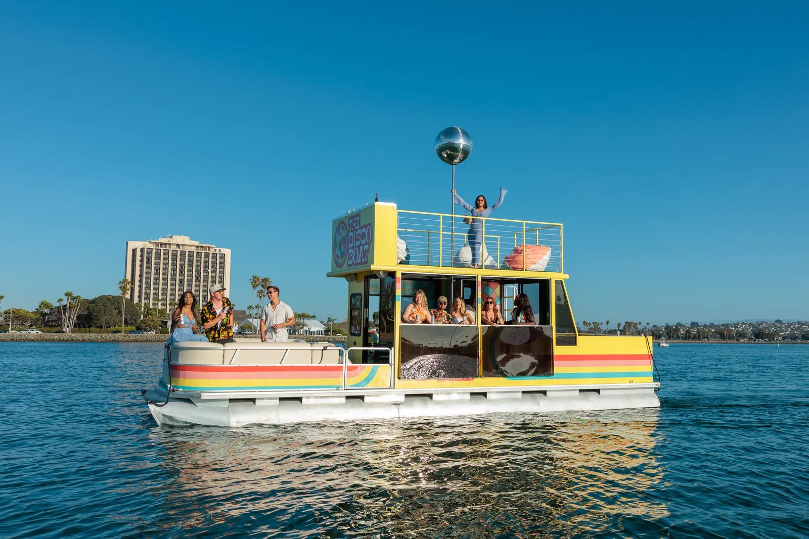 The Disco Boat at sunset on San Diego Bay — the rooftop mirror ball lit up, a guest waving from the upper deck, golden-hour light across the water