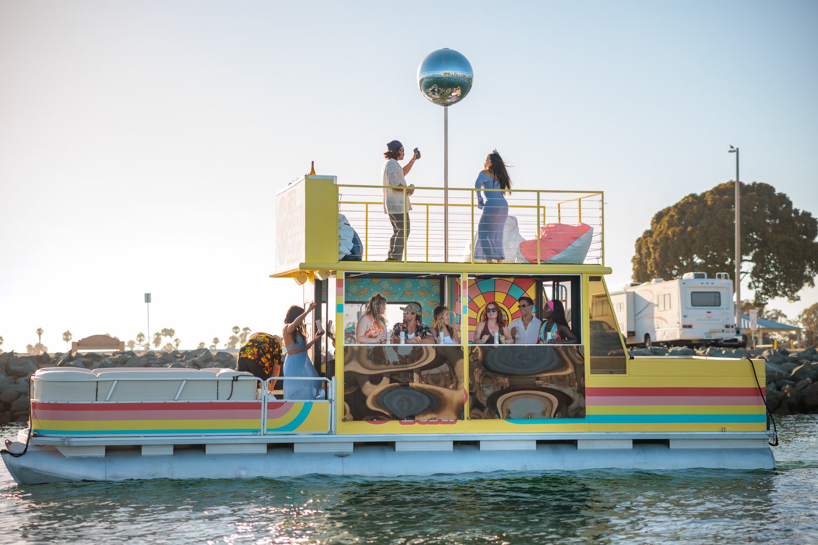 Themed cruise on the Disco Boat — guests on the rooftop with arms raised under the mirrored disco ball, side profile of the rainbow-striped hull on San Diego Bay